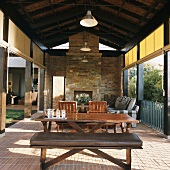 Wohn/Essraum im Freien - Terrasse mit rustikalem Kamin unter der offenen Dachkonstruktion und seitlichen Sonnenschutz-Rollos