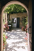 View through archway of sunny courtyard with shady, vine-covered pergola and abundant flowers