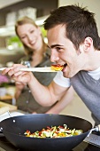 Young man tasting vegetables from wok, woman in background