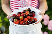 A child holding a white bowl of berries and cherries
