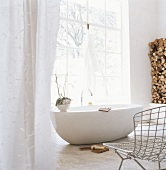 Eine runde freistehende Badewanne vor Panoramafenster im Badezimmer mit Drahtstuhl und gestapletes Brennholz in der Ecke