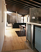 Blick von der Küche in einen offenen, modernen Wohnraum mit schräger Balkendecke, Betonwand und einem hellen Holzdielenboden