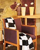 Black and white checked blanket on upholstered armchair with wooden frame; flowers on console table in background