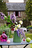 Kerzen & kleine Giesskanne mit Frühlingsblumen auf Gartentisch vor Haus