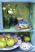 Coffee cup, flowers and bowl of apples on shelves