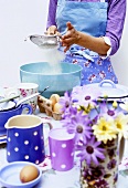 Woman sieving flour into a bowl, crockery & eggs in front