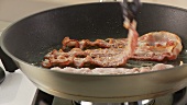 Rashers of bacon being fried and turned in a pan
