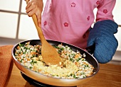Child Stirring Scrambled Eggs into a Wok Full of Rice Stir Fry, Wearing Oven Mitt While Holding Handle