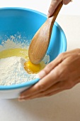Hand Mixing Egg into a Bowl of Flour with a Wooden Spoon
