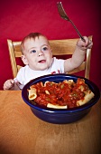 Baby at Table with Fork; Bowl of Pasta with Tomato Sauce on Table