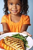 A young girl holding a plate of grilled chicken, grilled peaches and beans