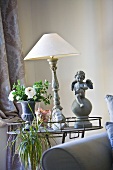 An antique table lamp with a white shade between a flower vase and an angel statue