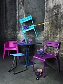 Mit Seilen aufgehängter, azurblauer Stuhl, darunter ein dunkelblauer Bistrotisch und violette Plastikstühle; dahinter eine graue, verwitterte Wand