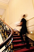 In einem eleganten Haus geht eine Person auf einem roten Teppichläufer die Treppe hinauf