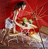 A side shot of a woman kneeling on a garden chair with a flower pot in her hand in front of a red sun umbrella