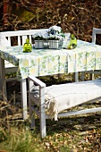 A white table and chairs with a floral patterned tablecloth and flowers in a garden