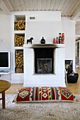 A floor cushion in front of a sooty fireplace and stack of firewood in a white stone shelf