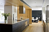 An open-plan kitchen - a shiny work surface and black drawers in front of a wooden partition wall and a view into a living room