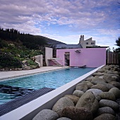 Langgestreckter Pool mit Findlingen vor Beckenrand und pinkfarbener Wandscheibe vor Haus