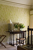 A bedroom with a table lamp on an antique wall table standing against a wall hung with floral wallpaper