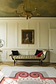 An elegant chaise longue in front of a white, wooden-panelled wall hung with a picture in an old-fashioned living room with a painted ceiling