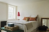 A white-painted bedroom with a stack of suitcases at the foot of a double bed and a bedside lamp with a red shade next to a window