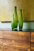 Green vases on a wooden side board in front of a yellow picture