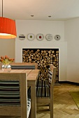 A dining table made of light wood with an orange lampshade in front of a fireplace stacked with wood