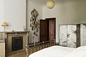 A bed room in a country house with a stainless steel bed and a painted paravent in front of a fireplace