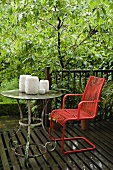 A red garden chair and a set of candles on a vintage metal table on a wooden terrace