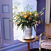 A bunch of flowers featuring yellow roses on a delicate wooden occasional table in front of lilac-painted window shutters