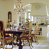 A laid table set with upholstered country house chairs and a chandelier with an open Mediterranean-style kitchen in the background