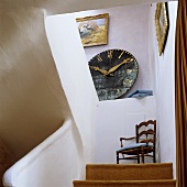 A flight of stairs with a view of a wall clock on a shelf and a wooden chair on the landing in the stairwell of a country house