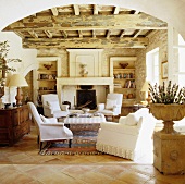 Blick durch Rundbogen in Mediterraner Wohnraum mit rustikaler Holzdecke und weiße Sesseln vor Kamin in Natursteinwand