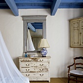 An old fashioned chest of drawers in front of a framed mirror in a room with a blue wooden ceiling