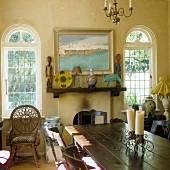 A dining table in front of a fireplace with African art on the mantelpiece and windows with round arches in a country house-style living room