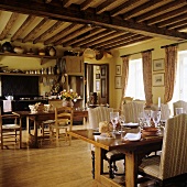 A set table with upholstered chairs in an open plan kitchen under rustic timber beam ceiling of an old country home