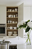 A view of a white crockery in a built-in cupboard with a bunch of flowers on a kitchen counter