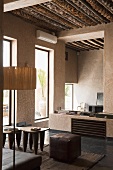 A room with a rustic wood beam ceiling with a leather stool and antique occasional tables in front of floor-to-ceiling windows in a Mediterranean house