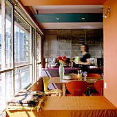 A view into an open-plan kitchen: a woman stands in the dining area with colored chairs in front of a glass facade
