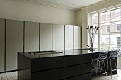 A black kitchen counter with bar stools in front of a window and a kitchen cupboard with a metal front