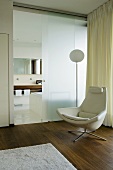 A room with walnut parquet and a designer leather armchair in front of an opaque glass wall with an open sliding door and view into a bathroom