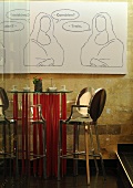 A designer bar table with red feet and a metal bar stools in front of a natural stone wall hung with a picture