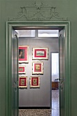 A view through a double door onto a collection of pictures hanging on the wall in red mounts