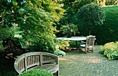 A garden - a wooden bench in front of a tree and a table with a chair on a brick-paved terrace