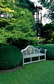 A white wooden bench in a niche between two box hedges and a view of a house with a balcony