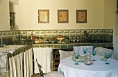 A country house kitchen - wine glasses on a table in front of a green-tiled kitchen counter and close curtains in front of cupboards