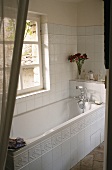 A view of a white tiled bathroom with a bathtub and a view of the garden
