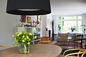 Black lampshade above flowers in a vase on a wooden dining table and an open plan living room