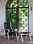 Sessel mit Fusshocker aus hellem Holzrahmen vor Gartentür aus Metall mit Ornamentmuster